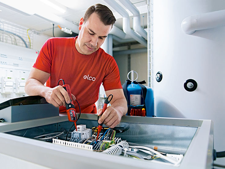 Servicetechniker arbeitet mit Werkzeugen an einer elektrischen Steuerung in einem Heizungs- oder Technikraum.